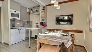 Eine kleine Küche mit Mikrowelle, Spüle und Schränken. Ein Esstisch mit Stühlen und ein an der Wand befestigtes Fernsehgerät.