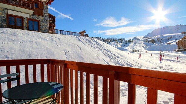Ein Holzbalkon mit einem Stuhl, der auf einen schneebedeckten Hang mit Skipisten und einem Berg im Hintergrund blickt.