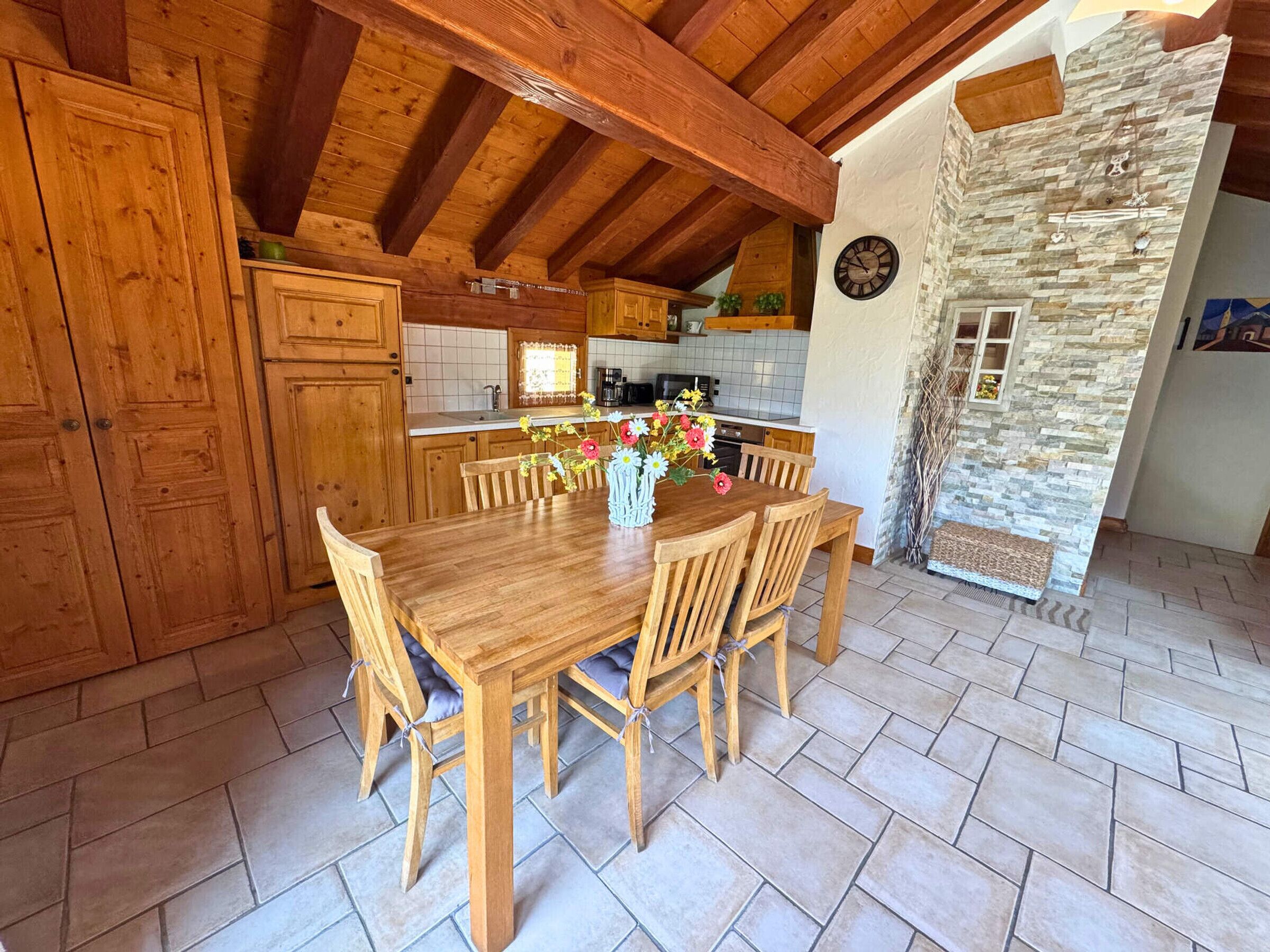 A wooden dining table with six chairs, a vase with flowers, and a kitchen in the background.