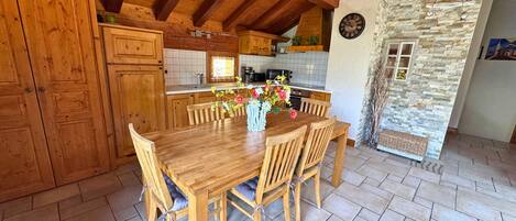 A wooden dining table with six chairs, a vase with flowers, and a kitchen in the background.