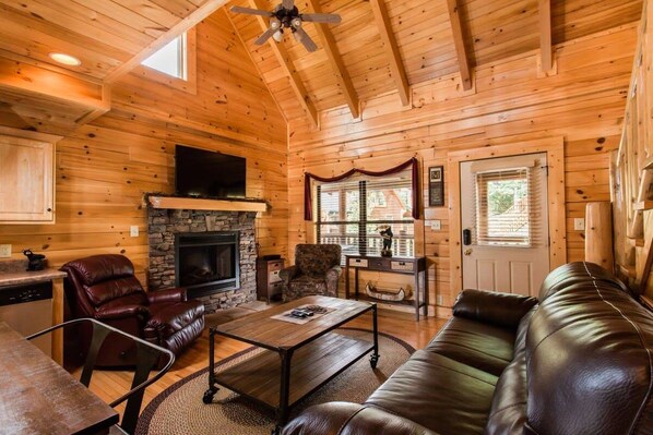 A log cabin interior with a stone fireplace, leather furniture, and a ceiling fan.