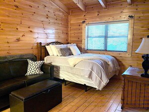 Habitación de cabaña de madera con cama, sofá, ventana con persianas y lámpara.