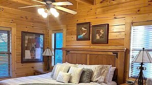 A wooden cabin bedroom with a bed, nightstand, lamp, ceiling fan, and framed pictures on the wall.