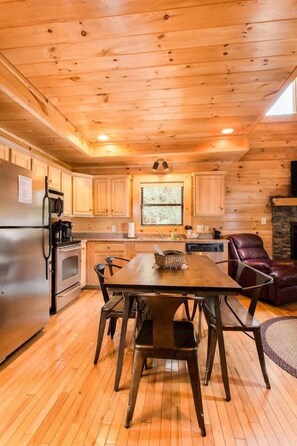 A wooden interior with a dining table, chairs, a refrigerator, and a stone fireplace. - 10 Min 2 Pigeon Forge Pkwy/Gorgeous Cabin/Hot Tub (Pigeon Forge)