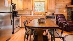 A wooden cabin interior with a dining table, chairs, a refrigerator, a stove, and a stone fireplace.