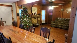 A wooden dining table with chairs, a Christmas tree, and a plaid sofa in a room with wooden walls and a ceiling fan. - Hideout in the Woods (Au Sable Charter Twp)