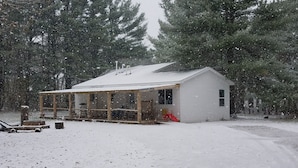 A small cabin with a covered porch, surrounded by snow-covered ground and trees. - Hideout in the Woods (Au Sable Charter Twp)