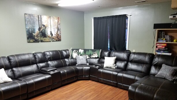 A living room with a L-shaped black leather sofa, wooden flooring, and two wall-mounted paintings. - Hideout in the Woods (Au Sable Charter Twp)