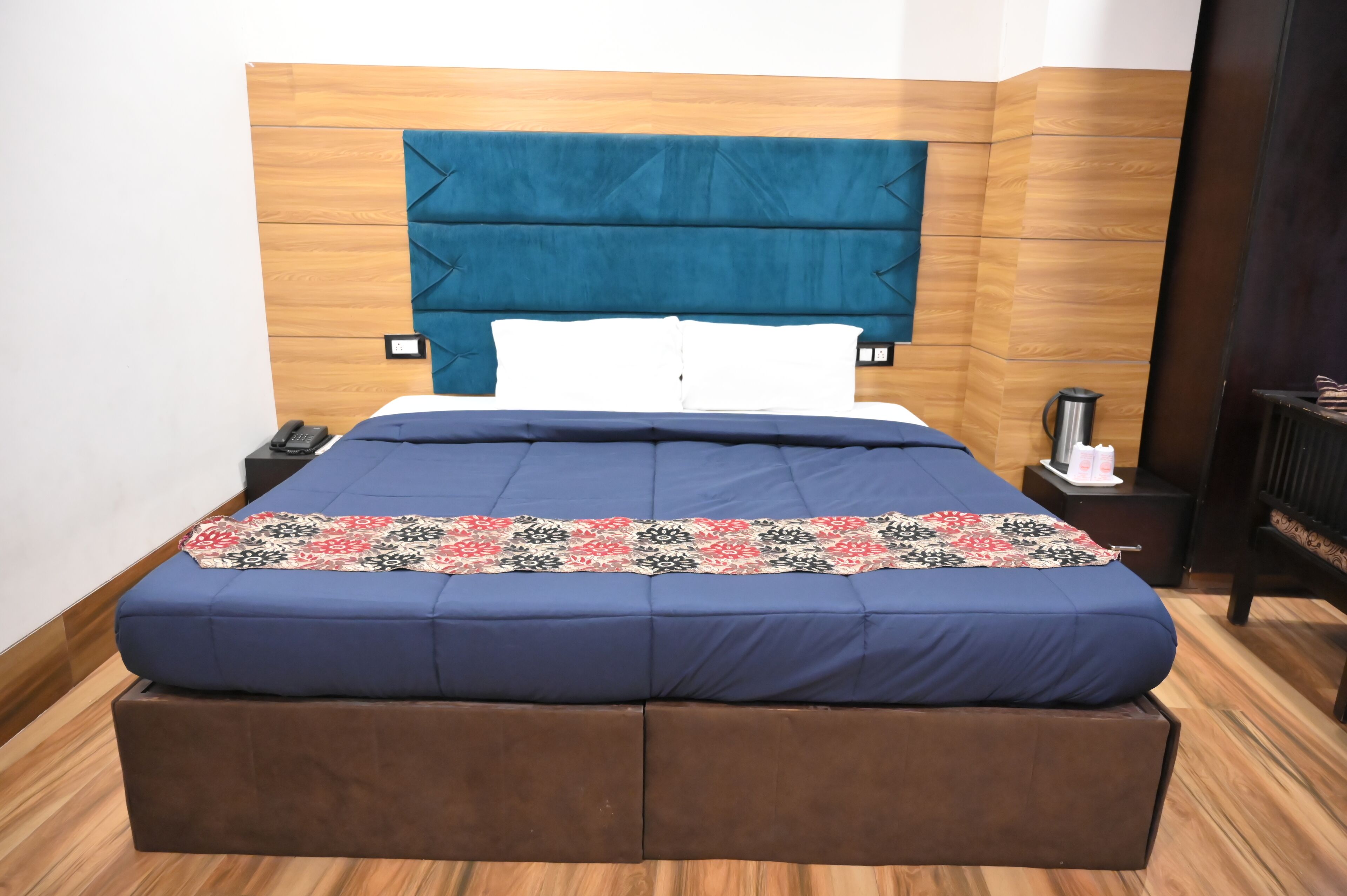Una cama con colchón azul y manta estampada, cabecera de madera y mesita de noche con teléfono y lámpara.