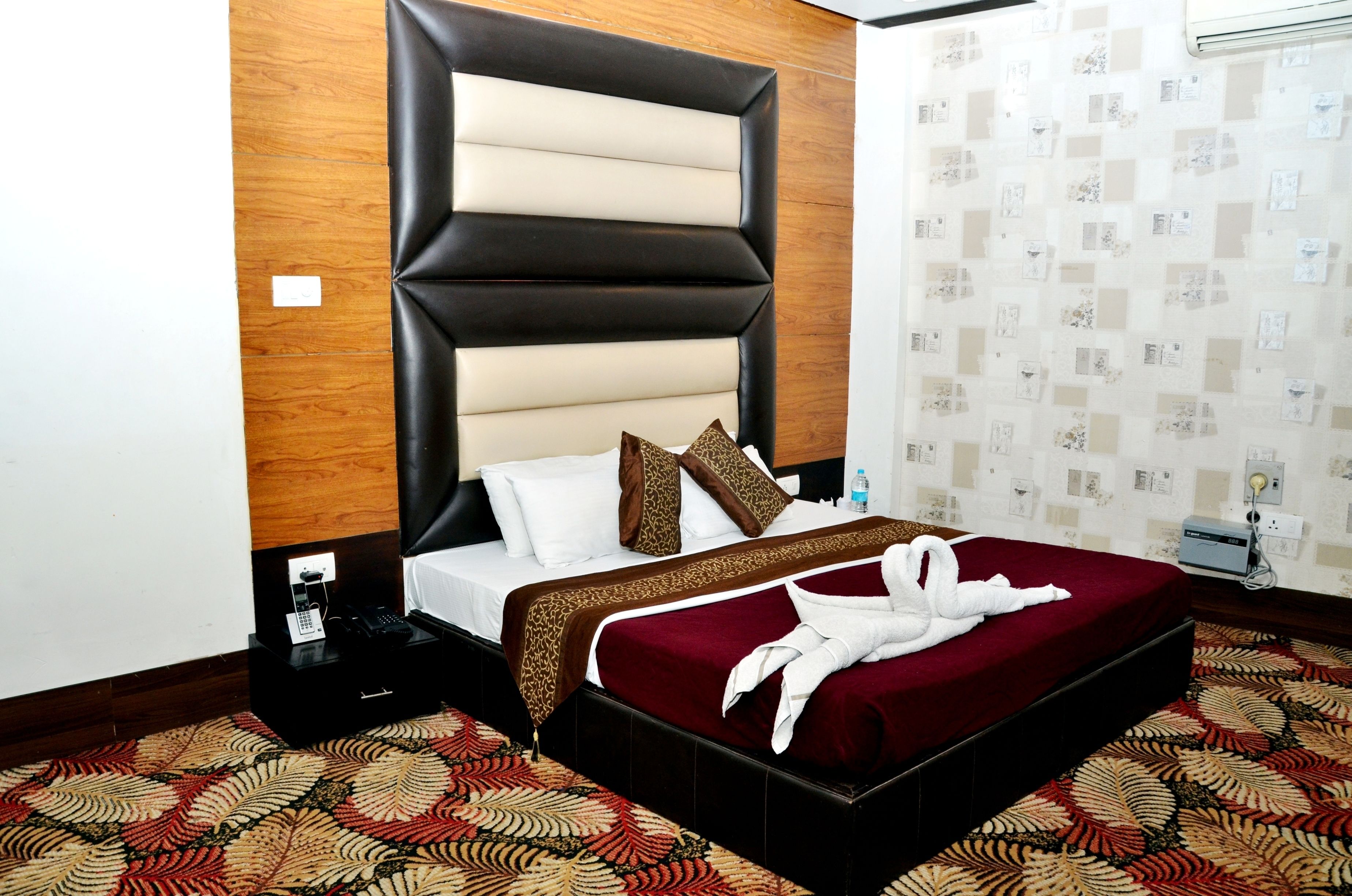A hotel room with a large bed, a wooden headboard, a patterned carpet, and a wall with a floral wallpaper design.