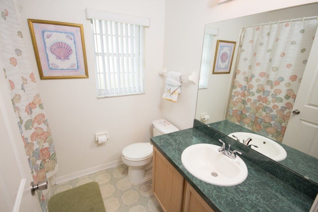 A bathroom with two sinks, a toilet, a window with blinds, a shower curtain with a shell pattern, and a framed picture on the wall.