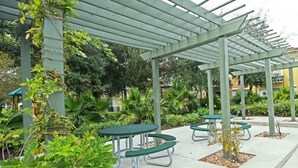 An outdoor patio area with green metal tables and benches, surrounded by lush greenery and a pergola structure. - Fv57771 - Emerald Island - 3 Bed 3 Baths Townhome (Kissimmee)