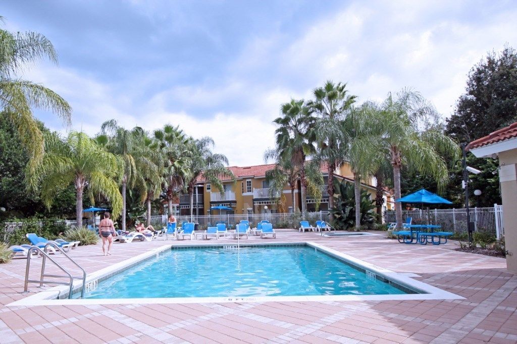 A swimming pool with lounge chairs and umbrellas, surrounded by palm trees and a building with a red roof.