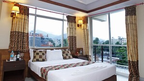 A spacious bedroom with a large bed, wooden headboard, and a view of the cityscape through large windows. - Durbar Suite (Pokhara)