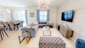 A modern living room with a grey sofa, a coffee table, a television, and a dining area with chairs and a table. - Aco237972 - Sonoma Resort - 6 Bed 4 Baths Villa (Kissimmee)