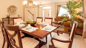 Une salle à manger avec une table en bois dressée pour quatre personnes, entourée de chaises. Un lustre se trouve au-dessus de la table, et un escalier mène à un autre niveau. On aperçoit un canapé et une plante en pot à l’arrière-plan.