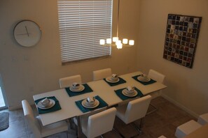 A dining area with a long table set for six, white chairs, a clock on the wall, and a window with blinds. - Aco106374 - Bella Vida Resort - 4 Bed 3 Baths Townhouse (Kissimmee)