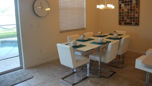 A dining area with a table set for four, white chairs, a clock, and a window with blinds. - Aco106374 - Bella Vida Resort - 4 Bed 3 Baths Townhouse (Kissimmee)