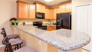 A kitchen with granite countertops, wooden cabinets, a black refrigerator, a microwave, and a stove. There are two bar stools and a small plant on the counter. - Fv49464 - Cypress Pointe - 5 Bed 4.5 Baths Villa (Davenport)