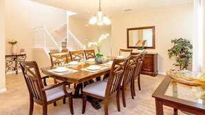 A dining room with a wooden table set for six, surrounded by chairs with cushioned seats. A chandelier hangs from the ceiling, and a staircase is visible in the background. - Fv49464 - Cypress Pointe - 5 Bed 4.5 Baths Villa (Davenport)