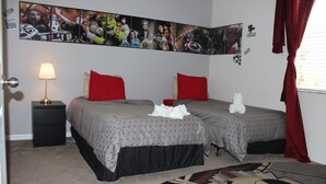 Ein Schlafzimmer mit zwei Betten, roten Kissen, einem Nachttisch mit Lampe und einem großen Wandbild mit Superhelden-Motiv.