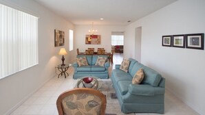 A living room with two teal sofas, a wooden armchair, a dining area with a chandelier, and a hallway leading to another room.