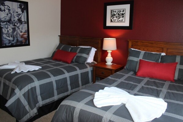 Habitación de hotel con dos camas, cada una con ropa de cama gris y roja, una lámpara blanca sobre una mesita de noche de madera, y un cuadro en la pared.