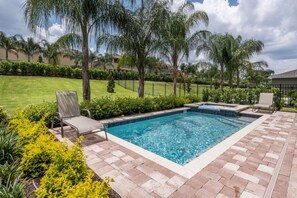 A rectangular swimming pool with a tiled edge, surrounded by a brick patio. There are two lounge chairs and a small hot tub area.