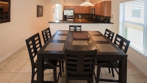 Un espace repas avec une longue table et des chaises, une cuisine à l’arrière-plan et une fenêtre avec des stores.