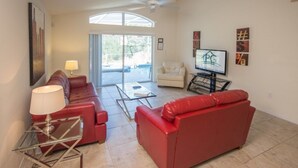 Un salon avec un canapé rouge, un fauteuil blanc, une table basse en verre, un téléviseur et un ventilateur de plafond.