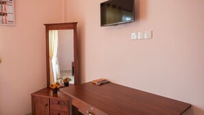 A bedroom with a wooden dresser, a mirror, a flat-screen TV mounted on the wall, and a small table with a book and a vase of flowers. - roylanka inn (Udunuwara)