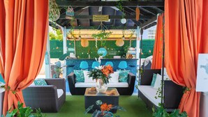 A covered outdoor seating area with wicker furniture, a glass coffee table, and orange curtains. - Hidden Gem Barbados (Inch Marlowe)