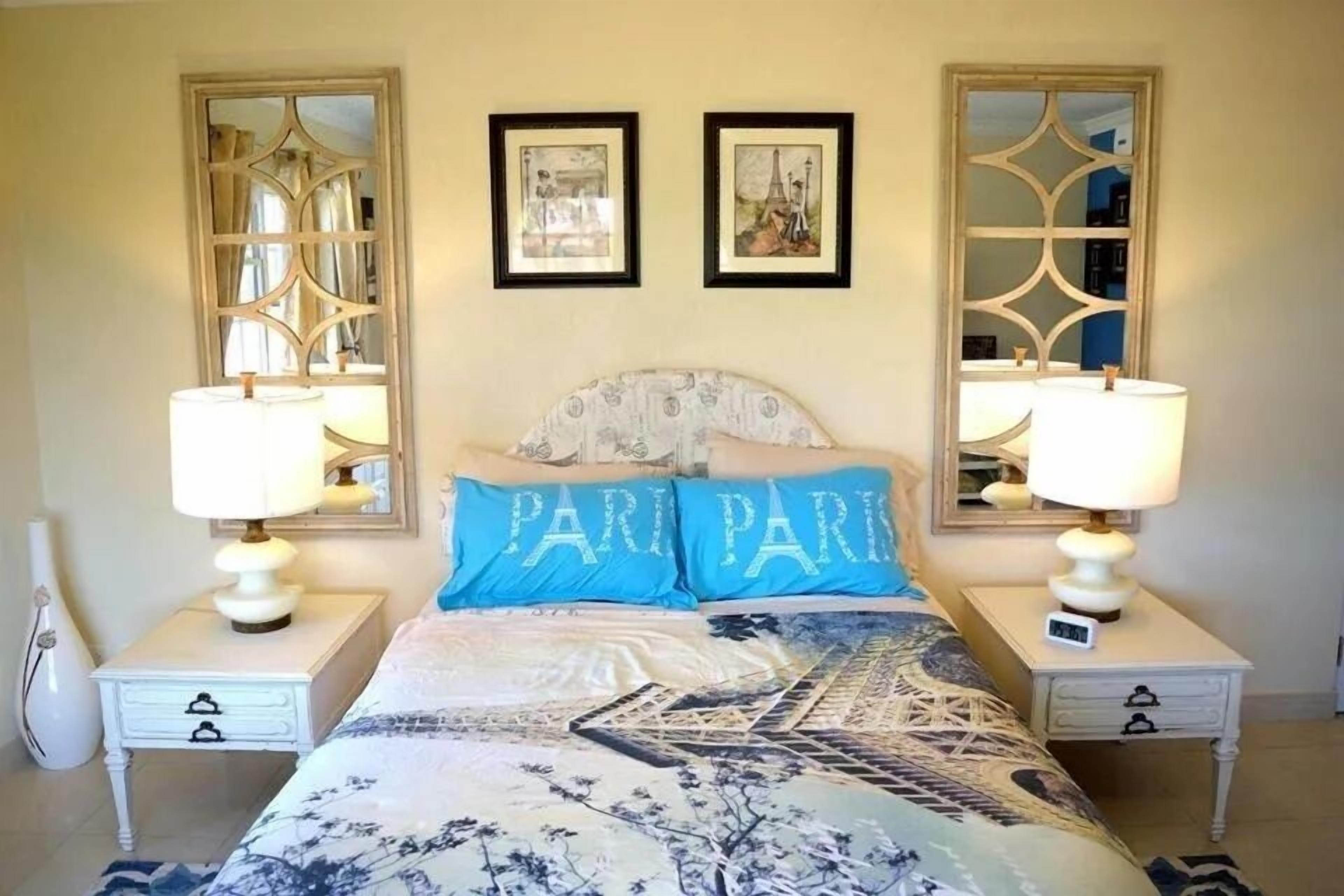 A bedroom with a bed, two bedside tables, two lamps, a mirror, and two framed pictures on the wall.