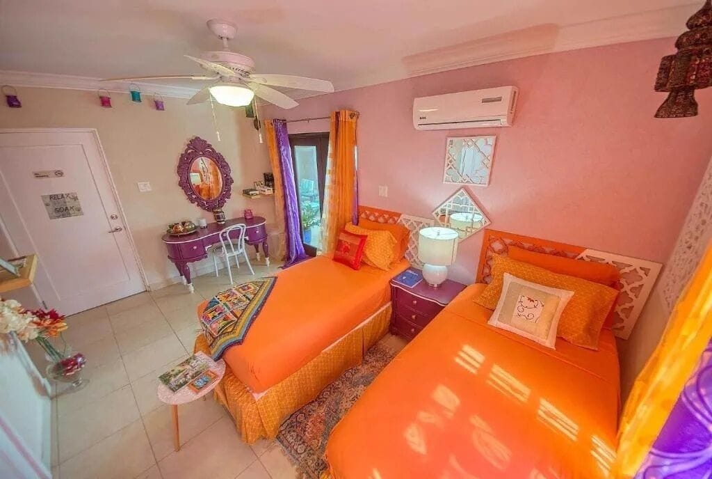 A room with two beds, a desk, a chair, a ceiling fan, an air conditioning unit, and a door with a sign.