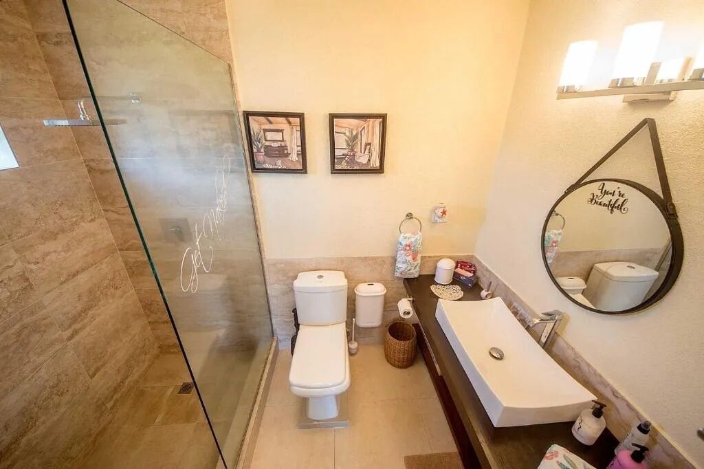 A bathroom with a glass shower enclosure, a toilet, a sink with a mirror, and two framed pictures on the wall.