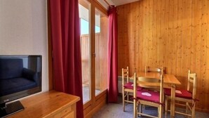 A room with a wooden wall, a wooden table with four chairs, a TV on a wooden stand, and a sliding glass door with red curtains. - Cozy studio for 4 at the foot of the slopes (Plagne 1800)