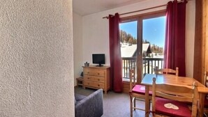 A room with a wooden table and chairs, a television on a wooden cabinet, and a view of a snowy landscape through a sliding glass door. - Cozy studio for 4 at the foot of the slopes (Plagne 1800)