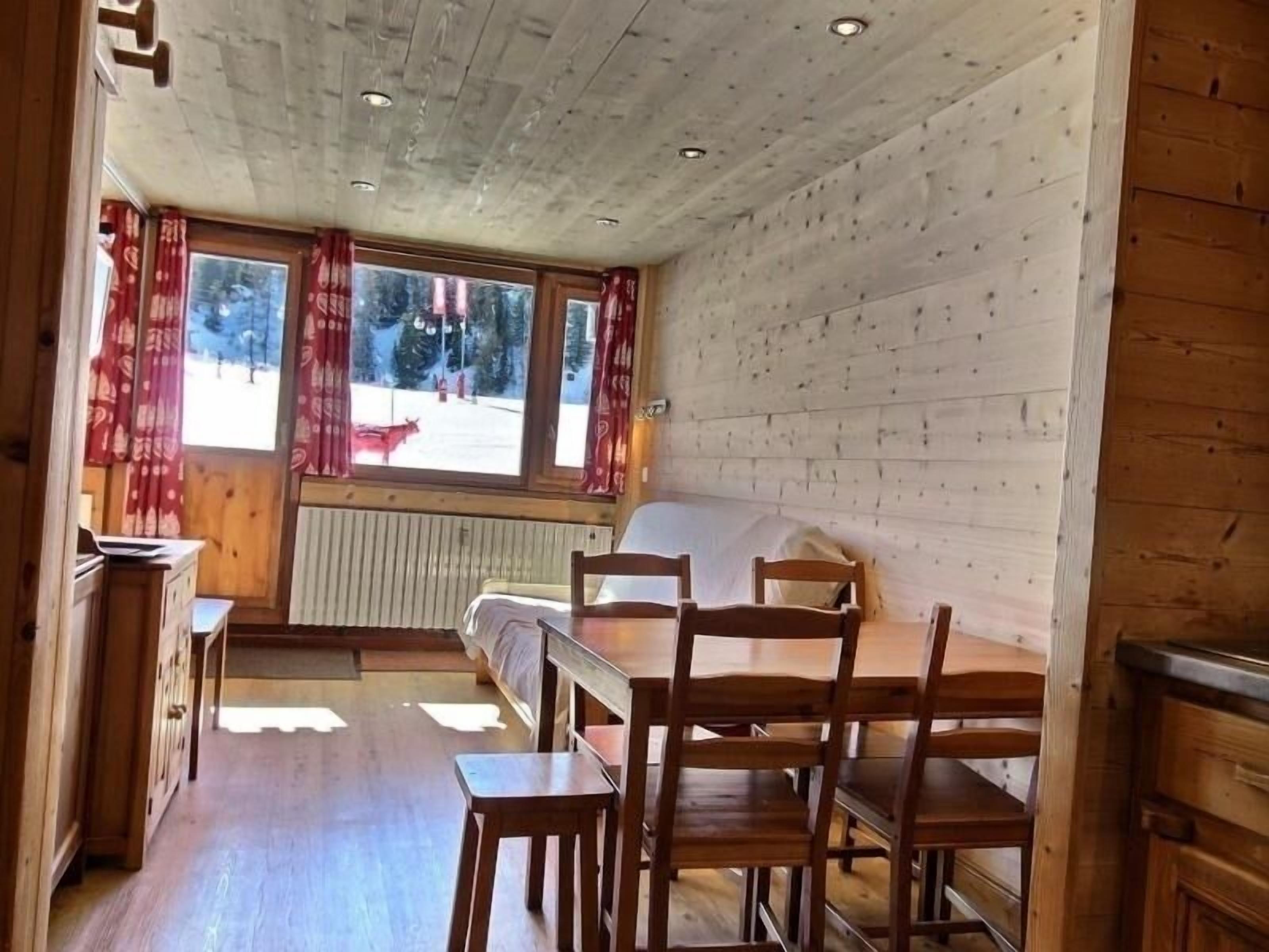 A cozy cabin room with a wooden interior, a bed, a dining table with chairs, and a view of a snowy landscape through the windows.