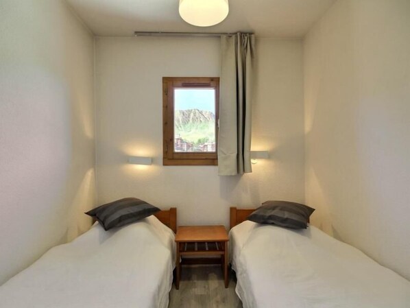 A small, clean room with two single beds, a wooden nightstand, a window with a view of mountains, and a ceiling light.