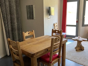 Uma mesa de jantar de madeira com quatro cadeiras, uma pequena mesa lateral de madeira, uma TV fixada na parede e uma porta com cortina vermelha.