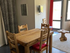 A wooden dining table with four chairs, a small wooden side table, a wall-mounted TV, and a door with a red curtain.