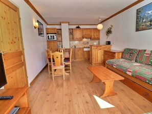 A cozy living room with a wooden sofa, a dining table, and chairs, a kitchen with wooden cabinets, and a television. - Cozy 4 Pax with Sunny Balcony (Méribel)