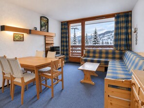 Ein Zimmer mit Esstisch, Stühlen, einer Couch und einem Fernseher. Ein Fenster bietet Blick auf schneebedeckte Berge.
