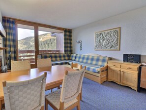 Un cuarto acogedor con una mesa de madera, sillas, un sofá azul y amarillo a cuadros, un aparador de madera y un ventanal con vistas al exterior.