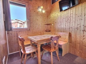 Un intérieur en bois avec une table et des bancs en bois, une fenêtre donnant sur des bâtiments et un téléviseur fixé au mur.