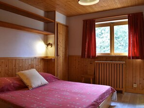 Et rum med træpaneler, en seng, et vindue med røde gardiner og en lampe monteret på væggen.