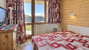 A bedroom with a wooden wall, a bed with a red and white quilt, a desk with a computer, and a window with curtains. - Apartment 6 people, south-west terrace at slopes' foot (Belle Plagne)