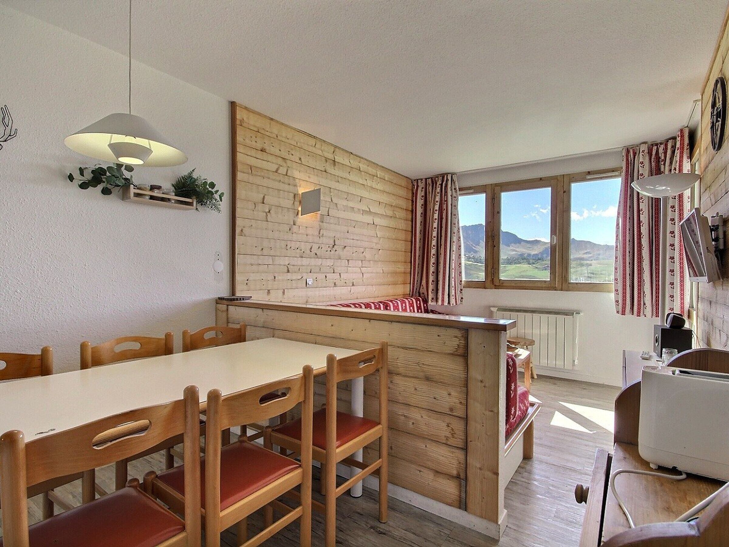 A cozy kitchen with a wooden wall, a dining table with chairs, a window with curtains, and a radiator.
