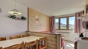 A cozy kitchen with a wooden wall, a dining table with chairs, a window with curtains, and a radiator. - Apartment 6 people, south-west terrace at slopes' foot (Belle Plagne)