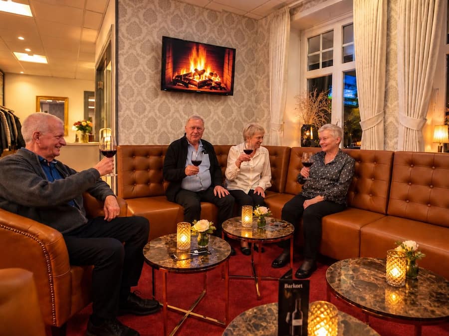 A group of people seated on a brown leather couch in a warmly lit room with a fireplace, a television mounted on the wall, and a small table with candles and a vase of flowers.